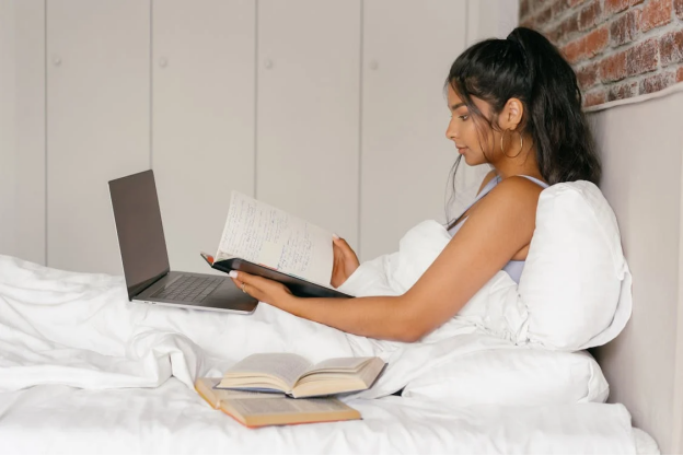 A student studying in bed with a laptop and books, showing a flexible approach to learning.