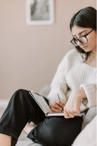 Young woman with glasses writing in a notebook, focused on personal and academic goals